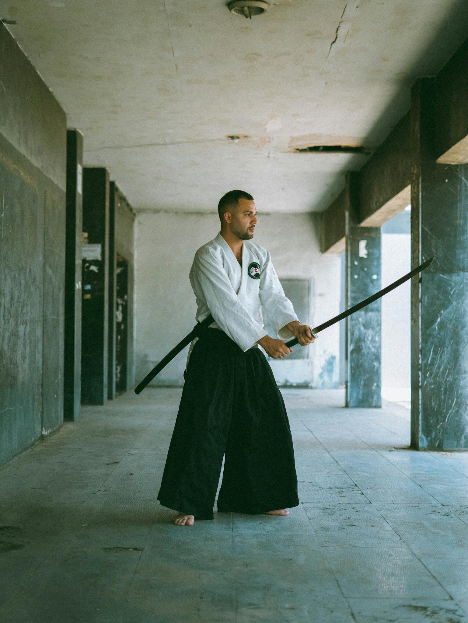 A man in traditional attire practices Kendo with a wooden sword in a spacious room.