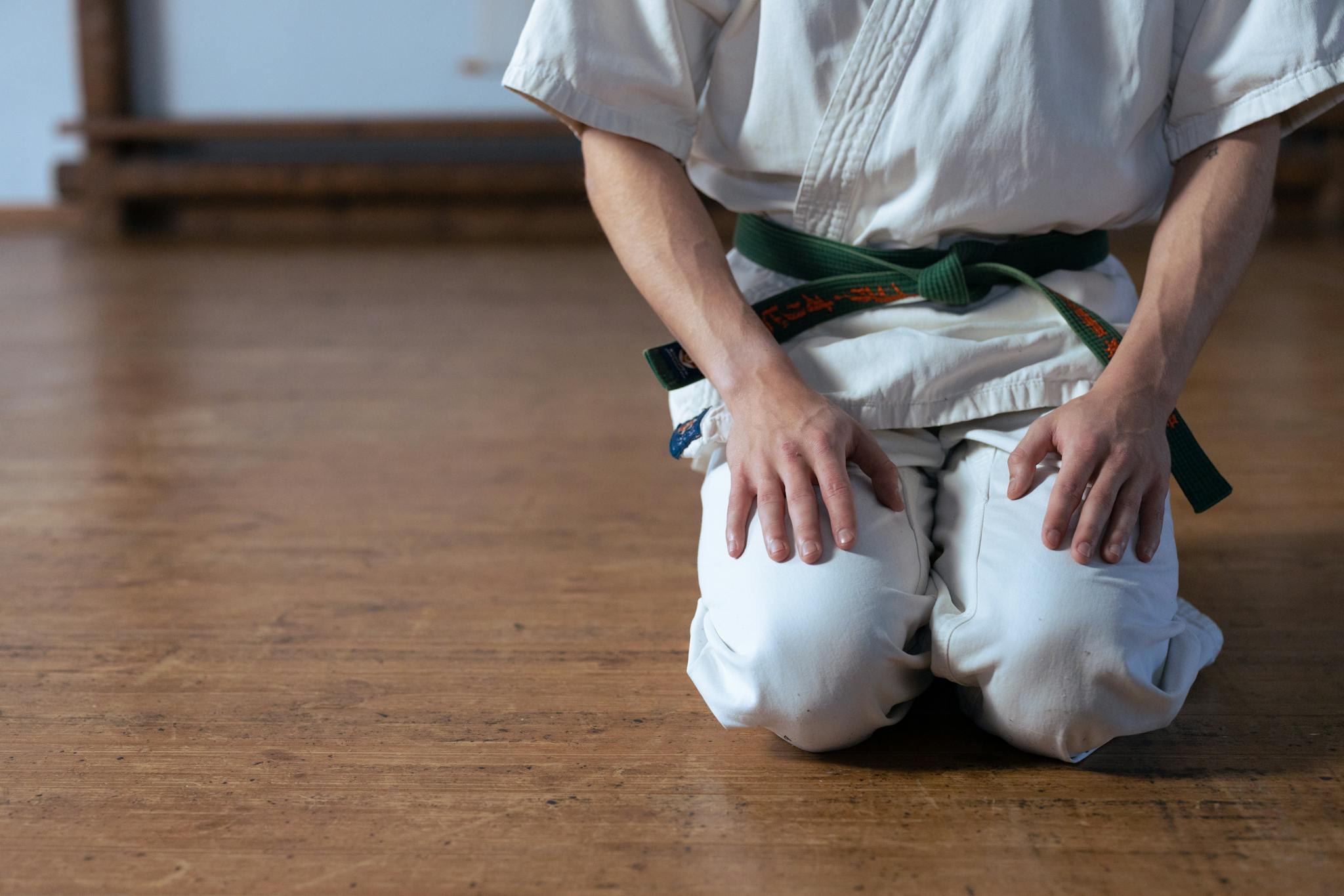 A martial artist in a white uniform kneels on a wooden dojo floor, focusing on practice.