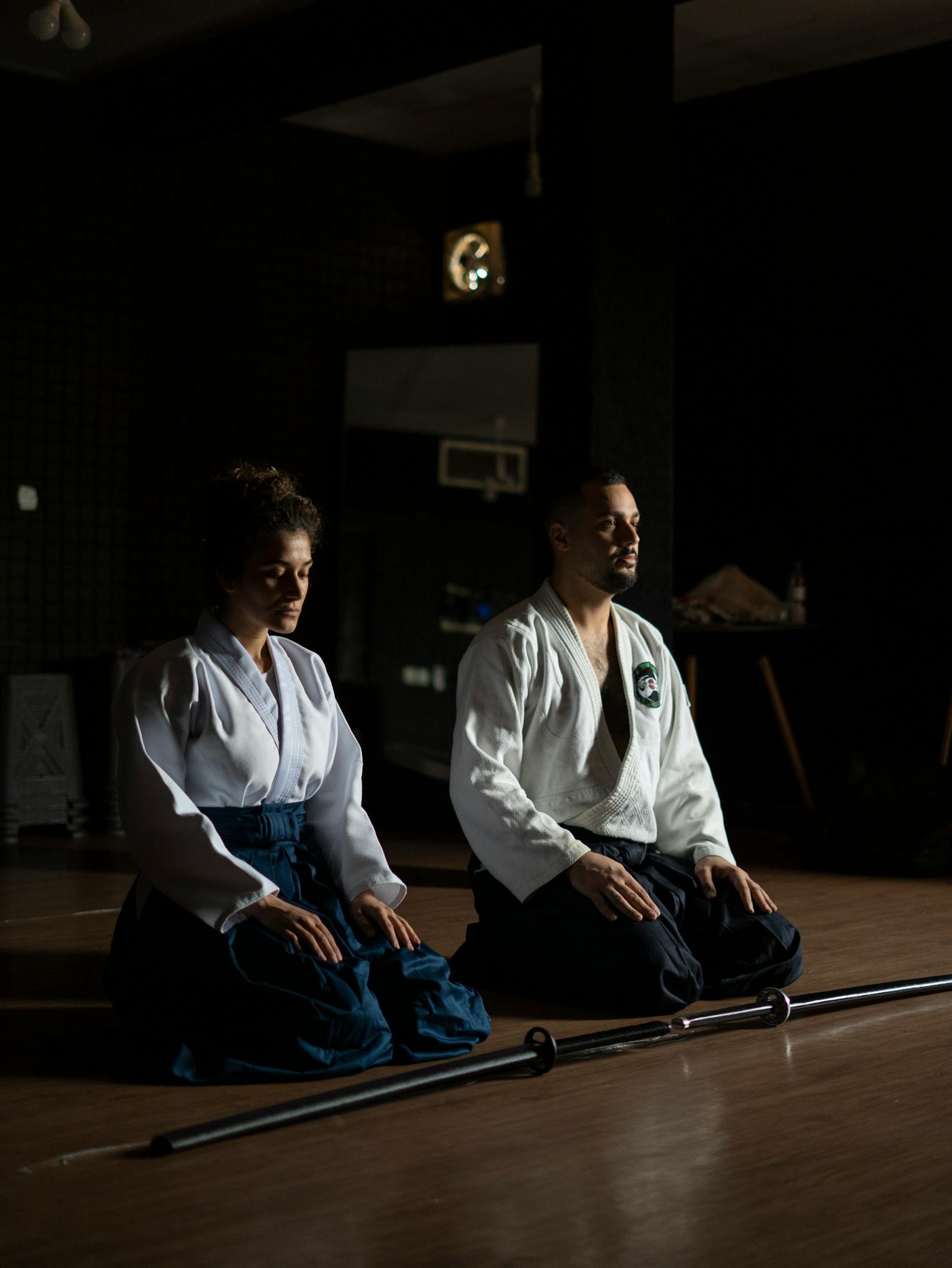 Two martial artists practice meditation in a dimly lit dojo, focusing on inner peace.